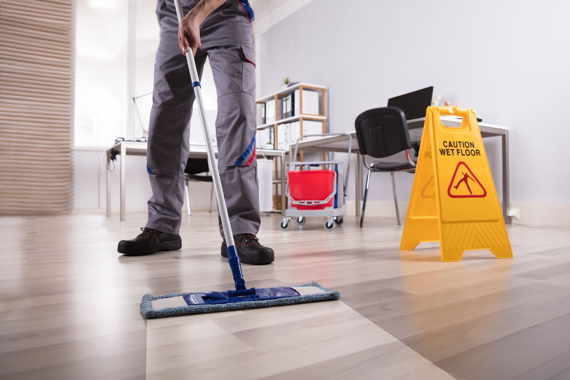 Eine Person wischt den Laminat-Fußboden eines Büros mit einem Mopp, daneben steht ein Warnschild 'Caution wet floor'